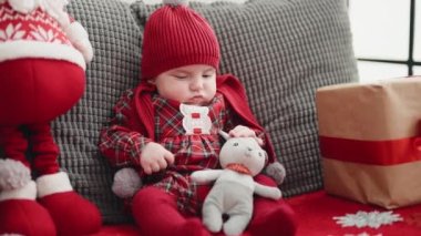 Sevimli beyaz bebek, Noel Baba oyuncağı ve Noel hediyesiyle kanepede oturuyor.
