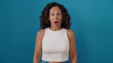 Middle age woman standing with surprise expression over isolated blue background