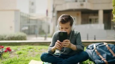 Sarışın çocuk parkta çimlerin üzerinde müzik dinliyor.