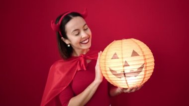 Young beautiful hispanic woman wearing devil costume holding halloween pumpkin lamp over isolated red background