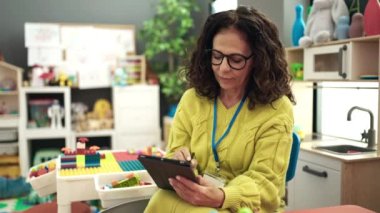 Middle age hispanic woman preschool teacher using touchpad at kindergarten