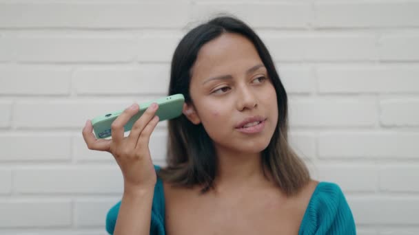 若いですヒスパニック女性笑顔自信に満ちたリスニング音声メッセージによってスマートフォン上の白い隔離されたレンガの背景 — ストック動画