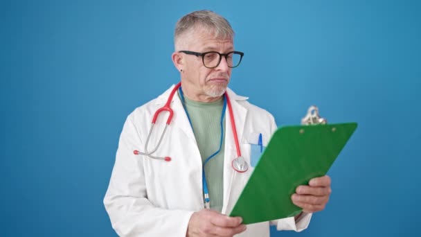 中年白发苍苍的男医生站在那里 带着轻松的表情阅读着孤立的蓝色背景的文件 — 图库视频影像