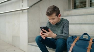 Okulda merdivenlerde oturan sarışın çocuk akıllı telefon kullanıyor.