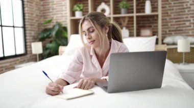 Young blonde woman using laptop writing on notebook at bedroom