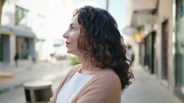 Orta yaşlı kadın gülümsüyor. Kollarını kavuşturup caddede duruyor.