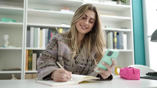 Young Blonde Woman Student Using Smartphone Writing Notebook Library University — Stock video