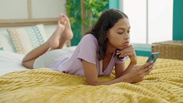 若いアフリカ系アメリカ人の女性がベッドルームでベッドの上に横たわって不幸な表情のスマートフォンを使用して — ストック動画