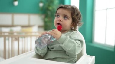 Adorable hispanic girl sitting on baby highchair drinking water at bedroom
