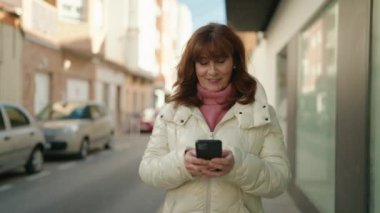 Orta yaşlı, kızıl saçlı, kendine güvenen bir kadın caddede akıllı telefon kullanıyor.