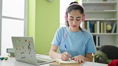 Laptop ve kulaklık kullanan güzel İspanyol kız öğrenci kütüphanede notlar alıyor.