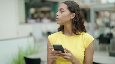 Young african american woman using smartphone with serious expression at coffee shop terrace