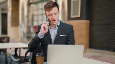 Kafkasyalı genç adam dizüstü bilgisayar kullanarak gülümsüyor ve kahve dükkanının terasında akıllı telefondan konuşuyor.