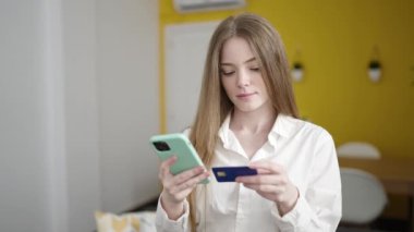 Young blonde woman shopping with smartphone and credit card thinking at home
