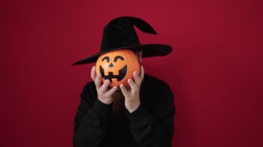 Young redhead man wearing wizard costume covering face with pumpkin basket over isolated red background