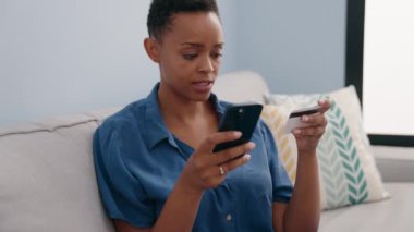 Akıllı telefon ve kredi kartı kullanan Afrikalı Amerikalı bir kadın evdeki kanepede oturuyor.