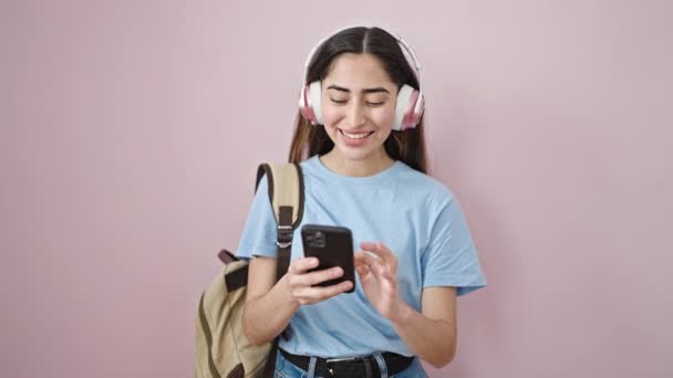 若いです美しいですヒスパニック女性学生使用スマートフォンとヘッドフォン上の隔離されたピンクの背景 — ストック動画