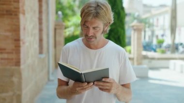 Young blond man smiling confident reading book at street