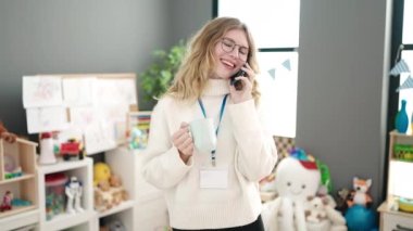 Young blonde woman preschool teacher talking on smartphone drinking coffee at kindergarten