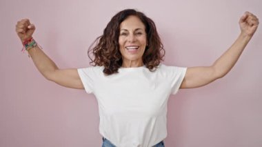 Orta yaşlı İspanyol kadın gülümsüyor, kendine güveni tam, kollarıyla soyutlanmış pembe arka planda güçlü hareketler yapıyor.
