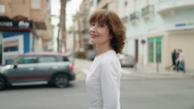 Orta yaşlı kadın gülümsüyor. Kollarını kavuşturup caddede duruyor.