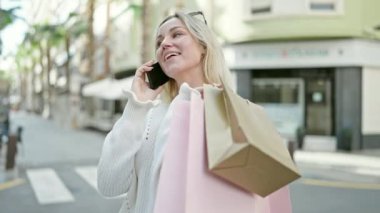 Genç sarışın kadın alışverişe gidiyor, elinde çantalarla caddede akıllı telefonla konuşuyor.