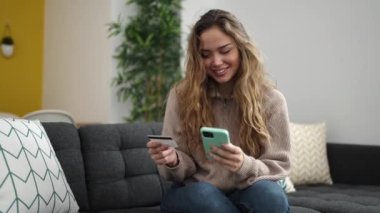 Akıllı telefon ve kredi kartı kullanan genç, güzel İspanyol kadın evdeki koltukta oturuyor.