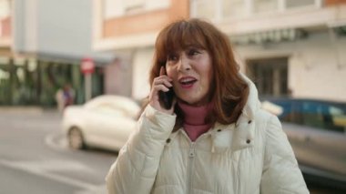 Middle age redhead woman smiling confident talking on the smartphone at street