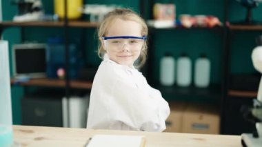 Sevimli sarışın kız öğrenci kendinden emin gülümsüyor. Laboratuvar sınıfında kollarını kavuşturup el kol hareketi yapıyor.