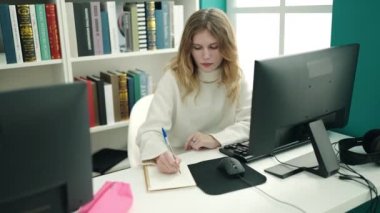 Young blonde woman student using computer writing on notebook at library university