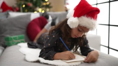 Adorable hispanic girl drawing on notebook lying on sofa by christmas tree at home