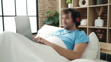 Young hispanic man using laptop and headphones lying on bed at bedroom