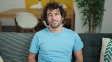 Young hispanic man holding blackboard with new home message sitting on sofa at home