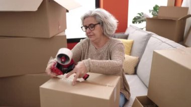 Middle age woman with grey hair smiling confident packing cardboard box at new home