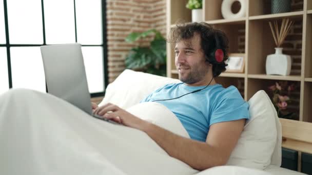 Young Hispanic Man Using Laptop Headphones Lying Bed Bedroom — Stockvideo