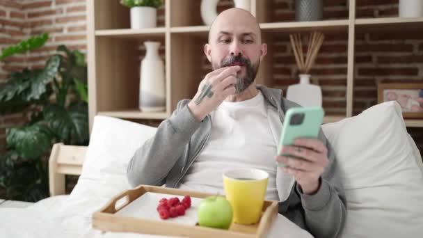 年轻的秃头男子在卧室用智能手机吃早餐 — 图库视频影像