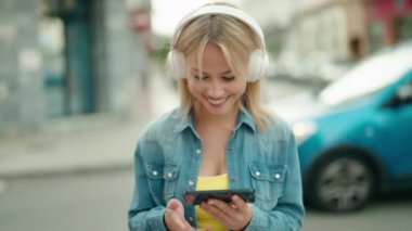 Genç sarışın kadın gülümsüyor. Sokakta akıllı telefondan videolar izliyor.