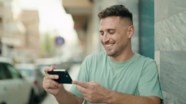 Genç İspanyol adam gülümsüyor. Sokakta akıllı telefondan videolar izliyor.