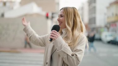 Young blonde woman journalist speaking using microphone at street