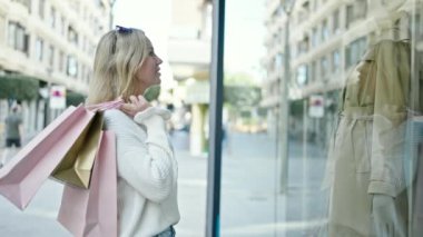 Gülümseyen genç sarışın kadın elinde çantalarla alışverişe çıkıp sokakta vitrine bakıyor.