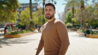 Genç İspanyol adam gülümsüyor. Parkta kollarını kavuşturmuş duruyor.