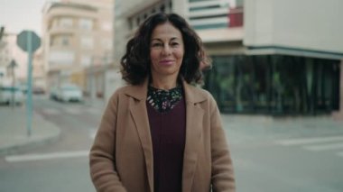 Orta yaşlı İspanyol kadın gülümsüyor. Kollarını kavuşturup caddede duruyor.