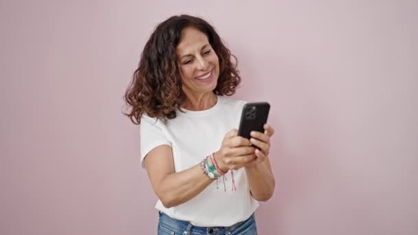 中年ヒスパニック女性使用スマートフォン笑顔上の隔離されたピンクの背景 — ストック動画