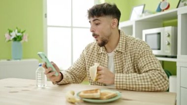 Genç Arap adam kahvaltısını evde akıllı telefon kullanarak yapıyor.