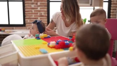 Öğretmen ve anaokulu öğrencileri anaokulundaki masada oturan inşaat bloklarıyla oynuyorlar.