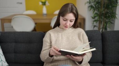 Young blonde woman reading book sitting on sofa at home