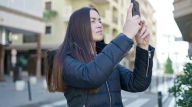 Beyaz bir kadın akıllı telefonla fotoğraf çekiyor. Sokakta ciddi bir yüz ifadesi var.