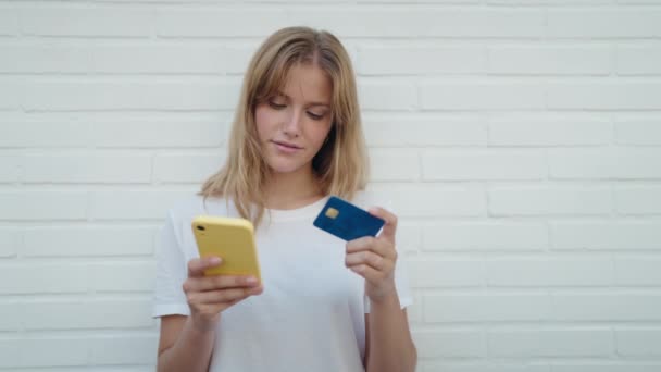 若いですブロンド女の子使用スマートフォンとクレジットカード上の隔離されたホワイトレンガの背景 — ストック動画