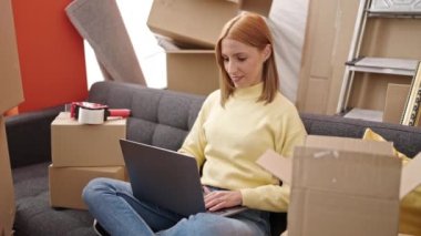 Young blonde woman using laptop sitting on sofa at new home