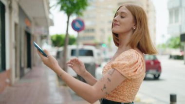 Genç kızıl saçlı kadın gülümsüyor. Kendine güveni tam. Sokakta akıllı telefonun yanında selfie çekiyor.